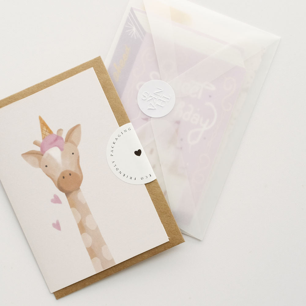 A baby giraffe unicorn greeting card with a kraft envelope, shown next to a clear paper packaging sleeve with a white sticker.