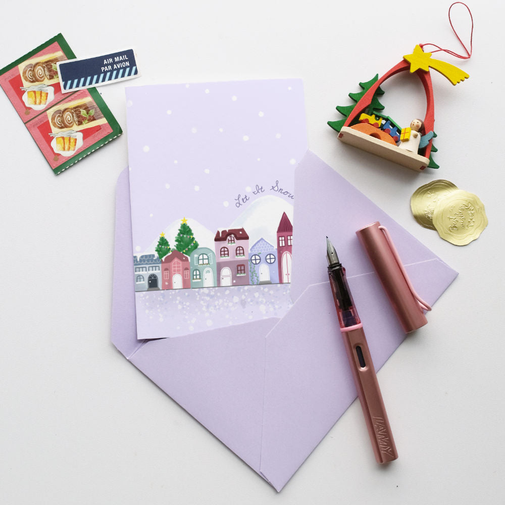 A styled flat-lay featuring a lavender greeting card with a winter village illustration and the words 'Let It Snow', accompanied by a matching lavender envelope, a pink fountain pen, and small holiday decorations on a white background.
