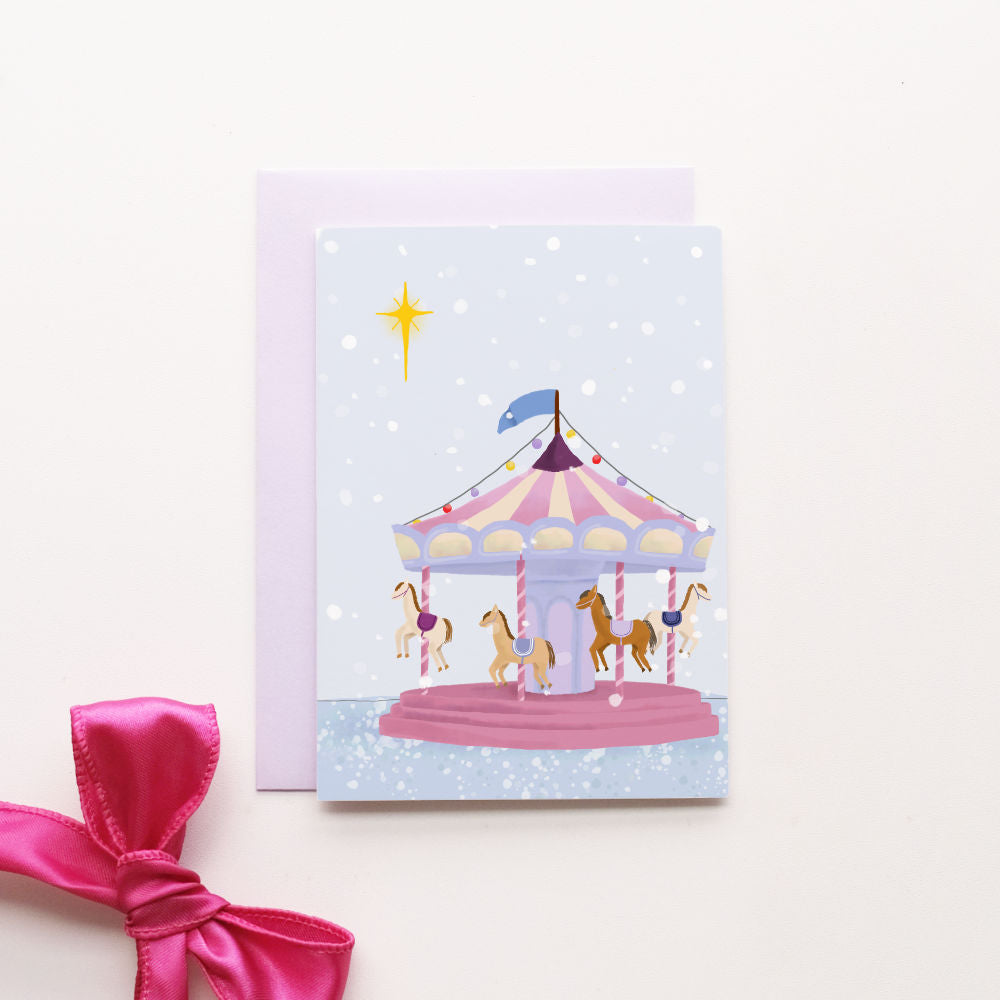 A flat-lay of a pastel purple envelope and matching card featuring an illustration of a carousel under a starry night sky. 
