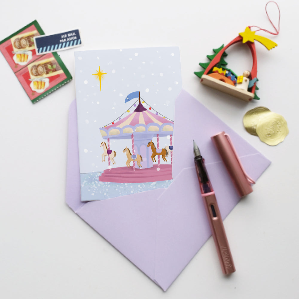 A flat-lay of a pastel purple envelope and matching card featuring an illustration of a carousel under a starry night sky. A rose gold fountain pen and pen cap sit beside them, along with small decorative props including a miniature Christmas tree, postage stamps.