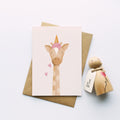 A cute greeting card featuring a giraffe wearing a pink ice cream, styled on a kraft paper envelope next to a wooden toy figure with a 'Believe My Love' tag, on a clean white background.