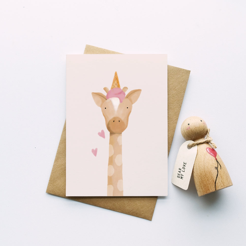 A cute greeting card featuring a giraffe wearing a pink ice cream, styled on a kraft paper envelope next to a wooden toy figure with a 'Believe My Love' tag, on a clean white background.