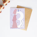 Greeting card with pink animal design and number 3, pen, and envelope on a white background