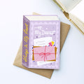 A decorative book-shaped greeting card or notebook with a lavender cover and spine, placed on top of a brown envelope on a white surface. The cover reads 'To: My Dearest' and shows an illustration of a pink gift box with a red bow and postmark.