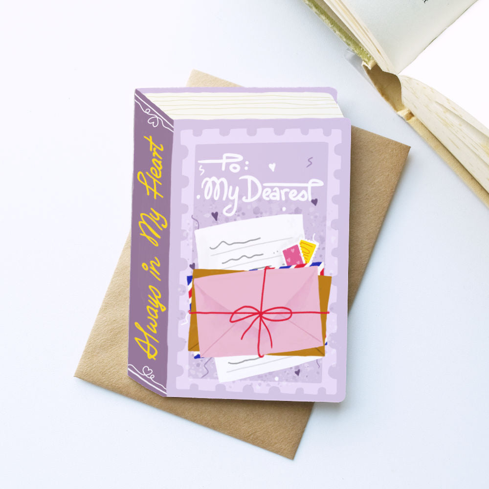 A decorative book-shaped greeting card or notebook with a lavender cover and spine, placed on top of a brown envelope on a white surface. The cover reads 'To: My Dearest' and shows an illustration of a pink gift box with a red bow and postmark.