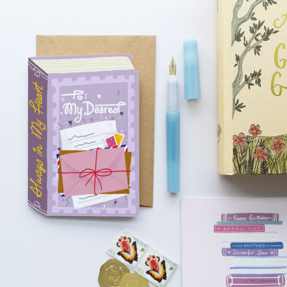 A flat-lay of stationery items on a white background, including a purple greeting card with an illustrated envelope and the words 'To: My Dearest', a kraft envelope, a light blue fountain pen, a floral notebook page, and small decorative stickers.