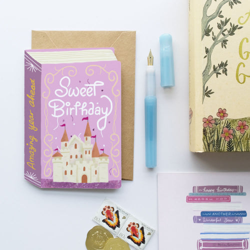 A styled flat-lay of a 'Sweet Birthday' book-themed greeting card with a brown envelope, a light blue fountain pen, a decorative book, stickers, and gold coins on a white background.