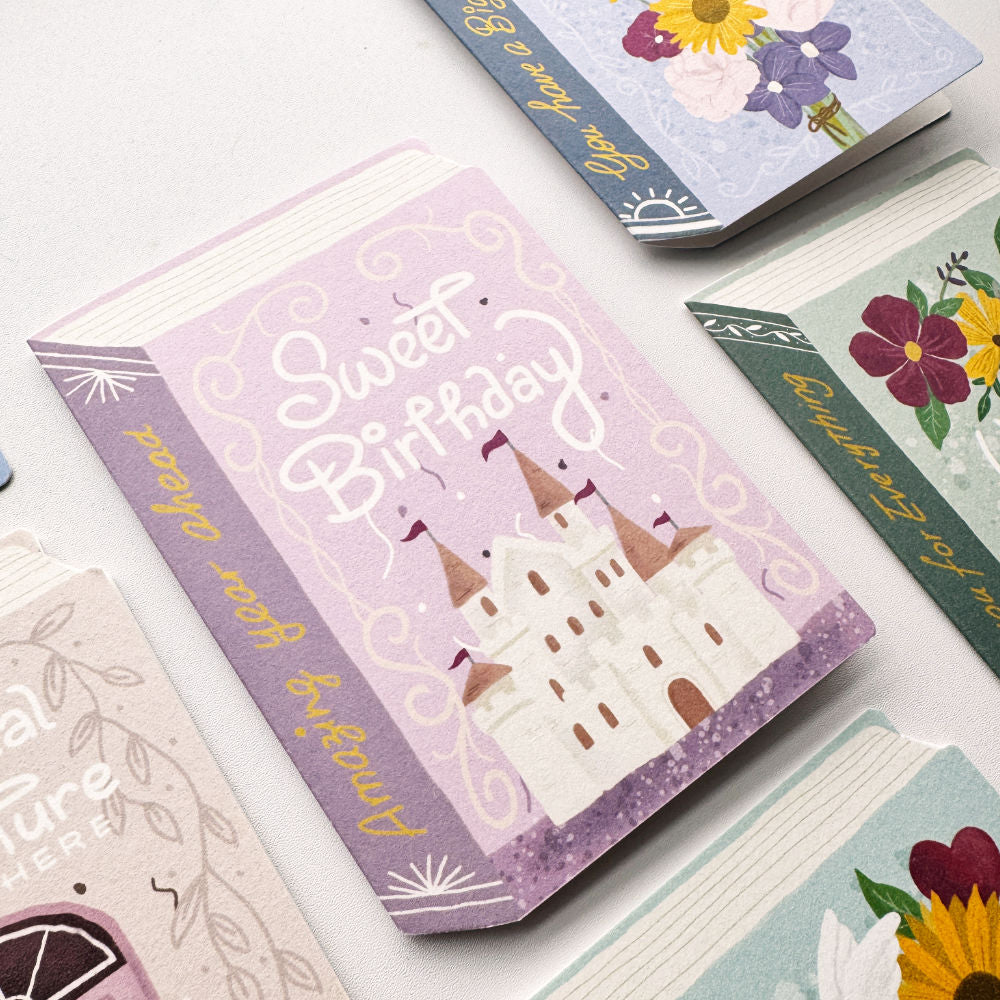 A stack of illustrated greeting cards arranged on a light surface. The top card is pink with a castle illustration and the words 'Sweet Birthday' in white script.
