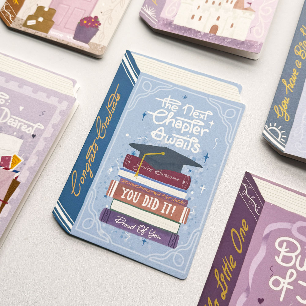 A close-up of a decorative book-shaped notebook or card with a light blue cover reading 'the Next Chapter Awaits' and 'YOU DID IT!' with a graduation cap illustration. Other similar stationery items are partially visible around it on a light surface.
