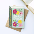 A book-shaped 'Words of Gratitude' thank you card styled on top of a brown kraft envelope, with an open book in the background on a white surface.