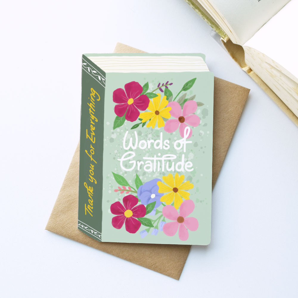 A book-shaped 'Words of Gratitude' thank you card styled on top of a brown kraft envelope, with an open book in the background on a white surface.