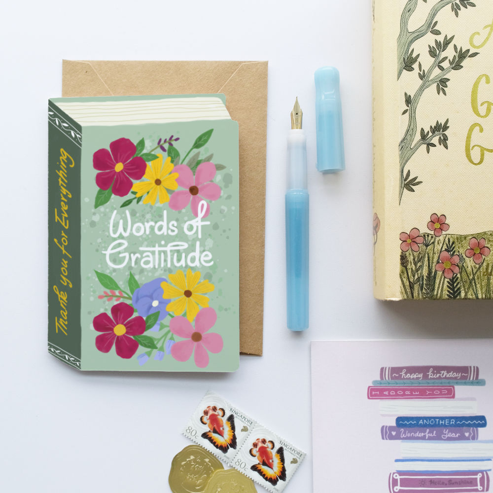 A flat-lay of a book-shaped 'Words of Gratitude' greeting card with a floral design, a kraft envelope, a light blue fountain pen, a decorative book, and small stickers on a white background.