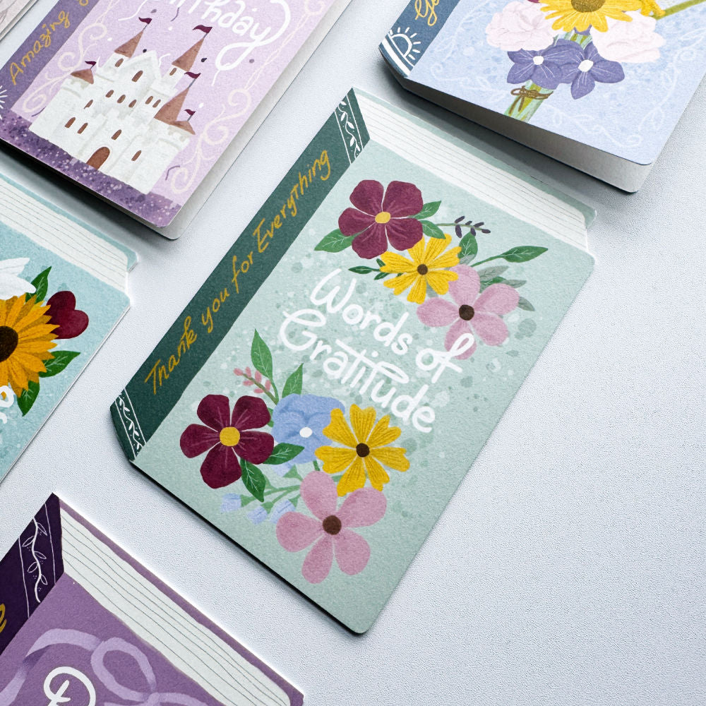 A stack of illustrated cards or booklets on a white surface, with the top card reading “Words of Gratitude” and decorated with colourful flowers. Other cards with pastel illustrations are partially visible around it.