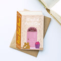 A decorative book-style card or notebook titled "Magical Adventure Starts Here!" with an illustrated pink door, standing on a brown envelope on a white surface. An open book is partially visible in the top-right corner.