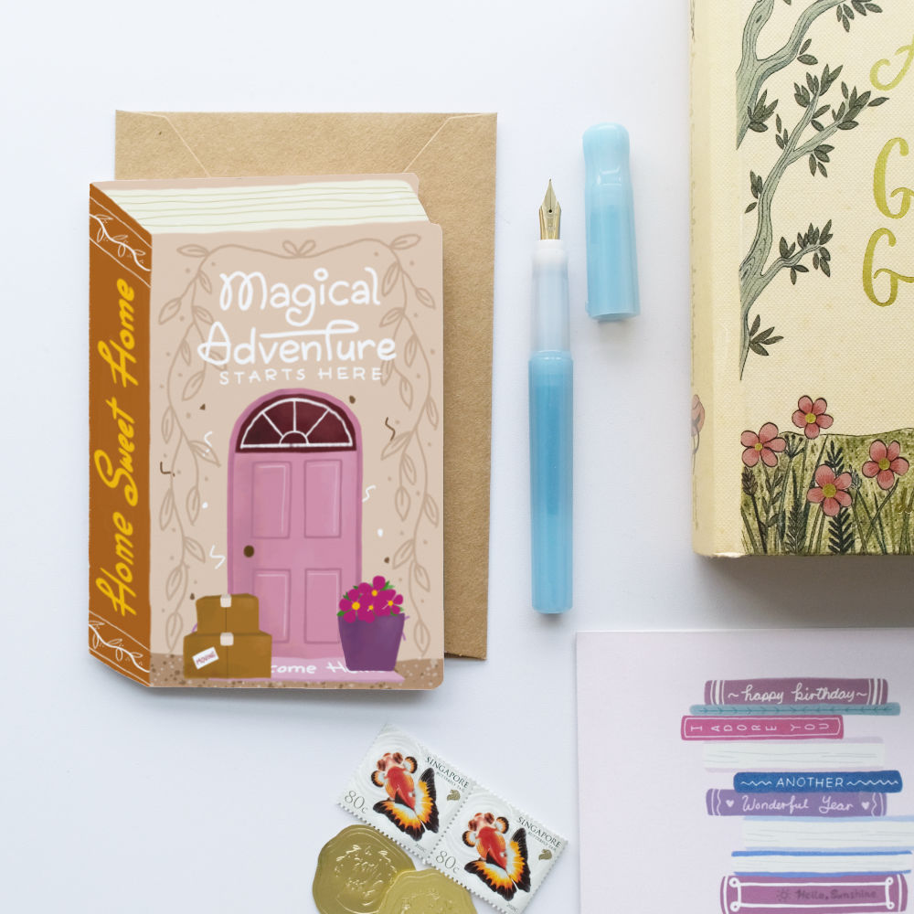 A styled flat-lay of stationery and gift items on a white background, including a small illustrated book-shaped card titled "Magical Adventure Starts Here!" with a pink door illustration, a kraft envelope, a light blue pen, part of a floral book or journal, and birthday sticker sheets.