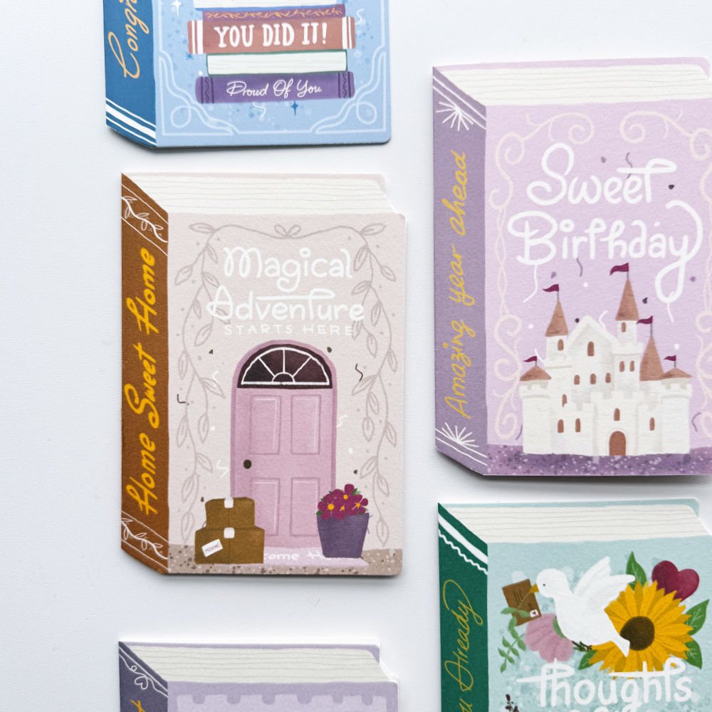 A set of illustrated greeting cards arranged on a white surface. The central card is a purple card titled "Bundle of Joy" with a baby stroller illustration; partial cards at the top and left show floral and text designs including "thoughts of you.