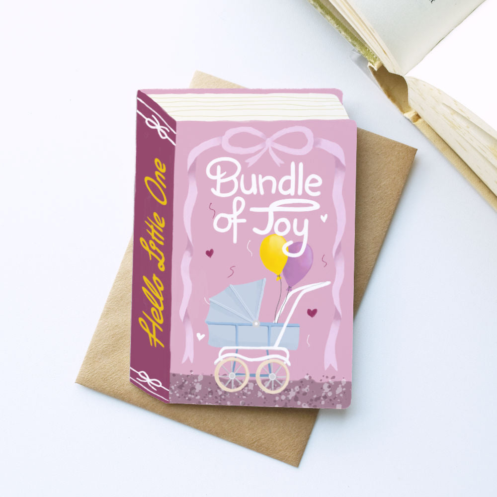 A baby-themed book-shaped greeting card set titled 'Bundle of Joy' featuring a pink cover with a baby stroller illustration and a brown kraft envelope, styled on a white surface with an open book in the background.