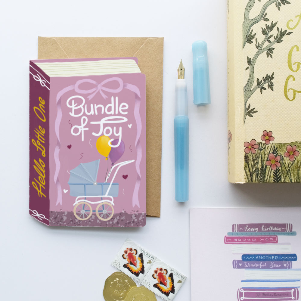 A styled flat-lay of a new baby card bundle on a white background. The set includes a pink book-shaped 'Bundle of Joy' baby carriage greeting card with a kraft envelope, a light blue fountain pen and cap, a gold foil 'Happy Birthday' book, a sheet of colorful stickers, and two gold coins.