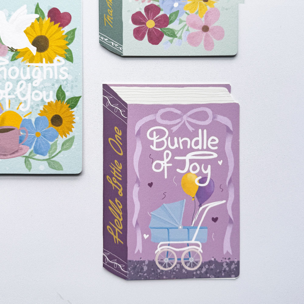 A set of illustrated greeting cards arranged on a white surface. The central card is a purple book-shaped card titled "Bundle of Joy" with a baby stroller illustration; partial cards at the top and left show floral and text designs including "thoughts of you.