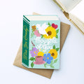 A book-shaped greeting card with a mint green cover and colourful floral illustration, placed on top of a brown envelope on a white surface. The card reads 'Thoughts of You' and 'Miss You Always'.