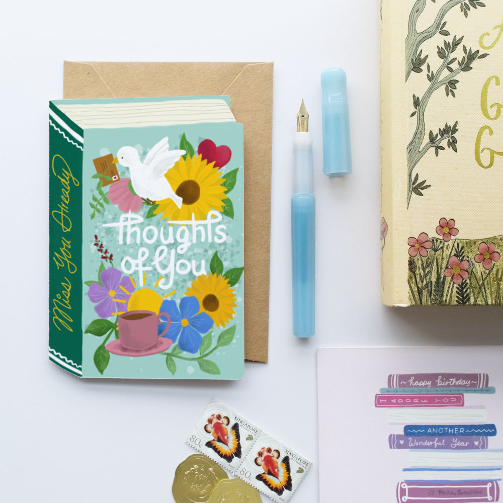 A styled flat-lay featuring a 'Thoughts of You' book-shaped greeting card with a green spine, a brown envelope, a light blue fountain pen, a blue pen cap, a gold foil stamp, and a decorative book on a white background.