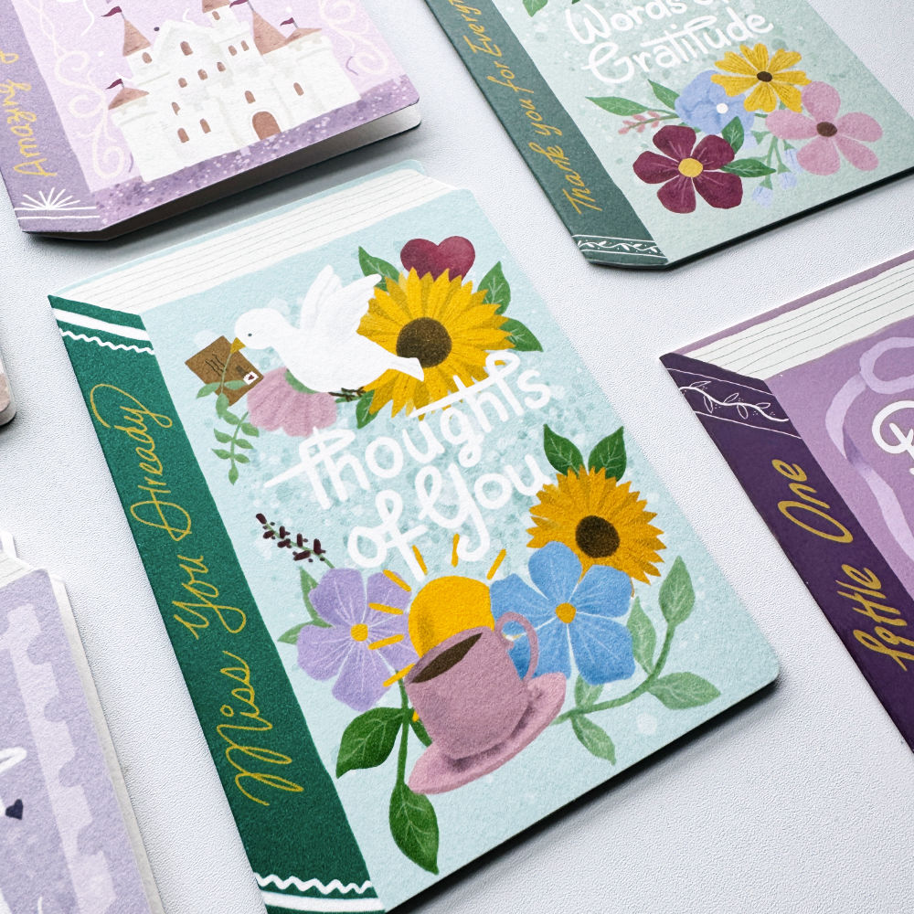 A book-shaped card with design featuring sunflowers, flowers, a white dove, a teacup, and the words 'Thoughts of You' on a light green background, styled on a white surface with other greeting cards partially visible around it.