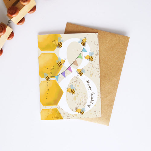 A honey-themed birthday card with a large white number 8 decorated with yellow honeycombs, bumblebees, and the words 'Happy Birthday', shown with a brown kraft envelope on a white background.