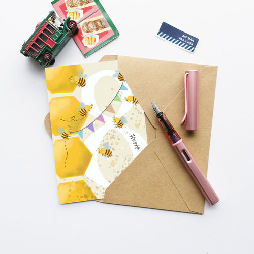 A styled flat-lay featuring a pink fountain pen and its cap on a brown kraft envelope, next to a honeycomb-patterned greeting card with bees and the word 'Honey'. A small toy truck, a postcard, and a sticker are arranged around it on a white background.