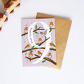 A  birthday card with a large white number 9, decorated with orange birds on brown branches against a pale pink background, shown with a brown envelope on a white surface.