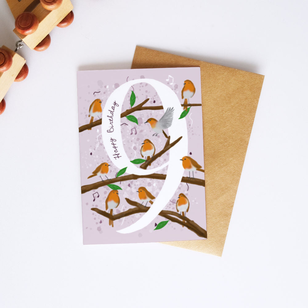 A  birthday card with a large white number 9, decorated with orange birds on brown branches against a pale pink background, shown with a brown envelope on a white surface.
