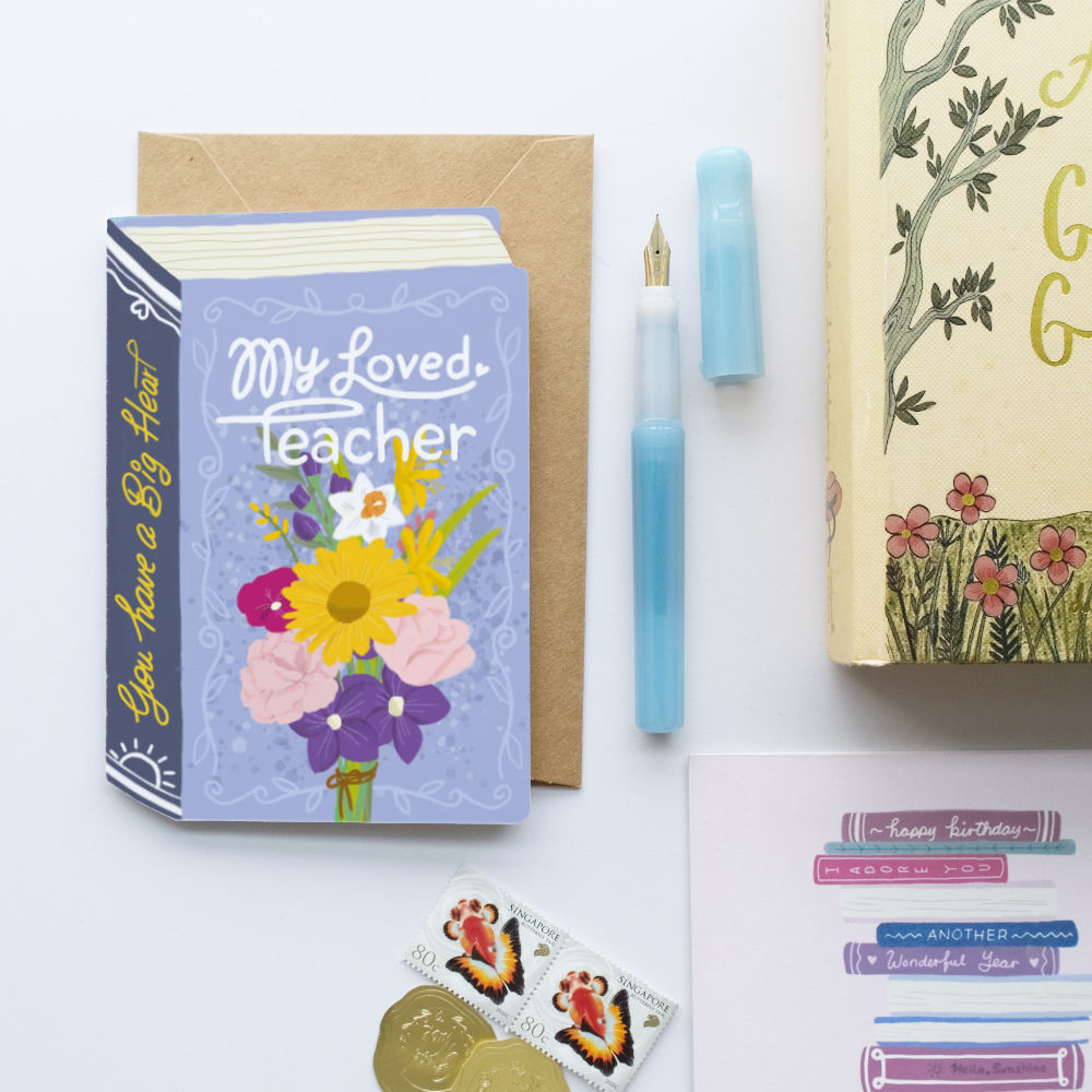 A styled flat-lay featuring a book-shaped greeting card with a lavender cover that reads 'My Beloved Teacher' and shows a bouquet of colourful flowers. The card is displayed with a brown kraft envelope, a light blue fountain pen, a matching pen cap, and a decorative book on a white background.