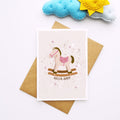 A baby greeting card featuring a cute rocking horse illustration with the text 'HELLO, BABY' and small heart accents, styled on top of a brown kraft envelope with a blue and yellow felt cloud decoration in the background.